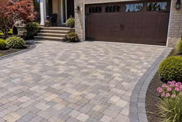 Residential paver driveway with interlocking gray and beige pavers, a stone home exterior, garage, and landscaped garden beds.