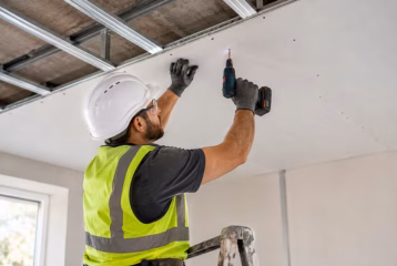 Contractor installing drywall ceiling panels with a drill on a residential construction project.