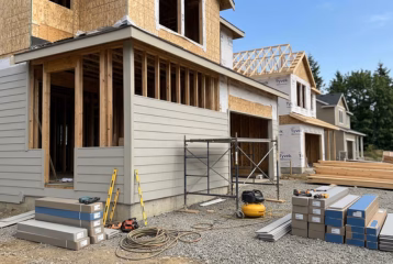 A detailed view of a two-story residential house under construction. The lower floor features installed light grey horizontal siding, while the second story shows exposed wood framing and OSB sheathing with prominent Tyvek house wrap visible on large sections. Scaffolding is set up on the second level. The gravel ground in the foreground is cluttered with construction materials, including stacks of packaged siding, a yellow air compressor, tools, and hoses. Other finished houses and a mature pine tree line are in the background under a blue sky.