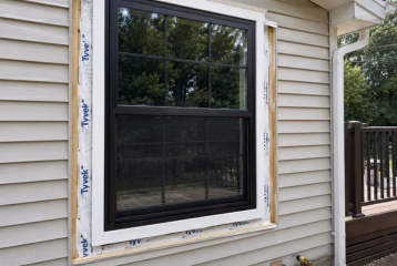 Residential window installation in progress with a new black-framed window, exposed flashing, exterior siding, and installation tools.
